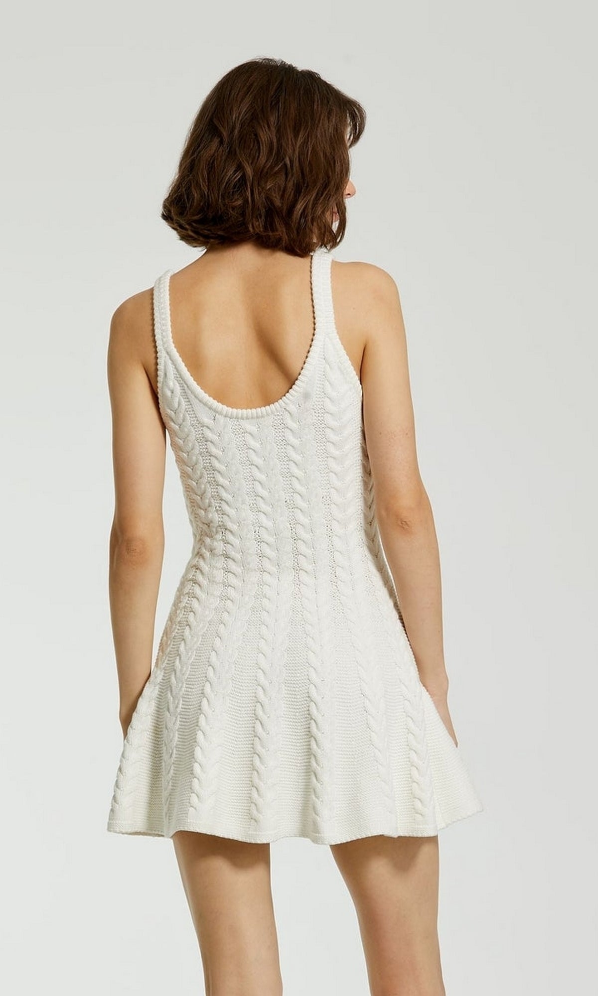 A woman with short brown hair wears the sleeveless, white Designer Short Knit Dress K12004 by Mac Duggal, featuring a chunky knit and open back, as she stands against a plain light background.